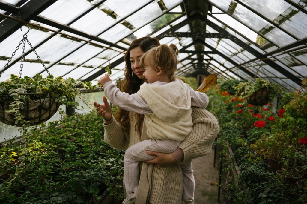 Smiling,Mother,Carrying,Daughter,Touching,Plant,In,Greenhouse