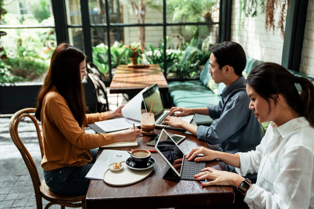 Three,Young,Business,People,Sitting,At,Table,,Using,Laptop,,Tablet,